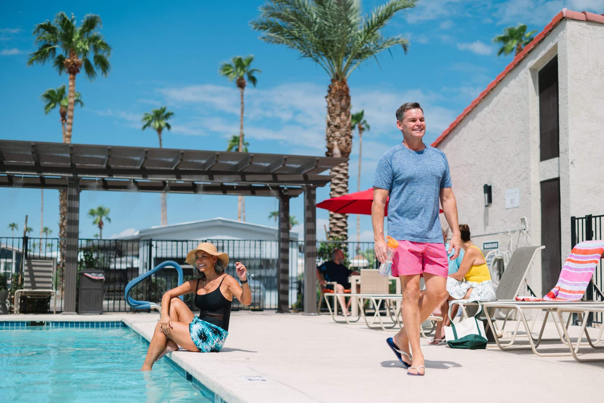 Outdoor poolside scene with palm trees, featuring a woman in a sun hat sitting by the water and a man in swimwear walking past. Other people relax in the background in Mesa, AZ